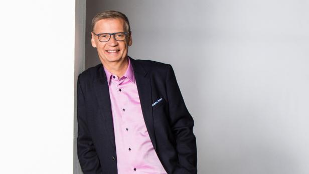 Günther Jauch lächelt in einem dunklen Sakko und einem hellrosa Hemd vor einem hellen Hintergrund.
