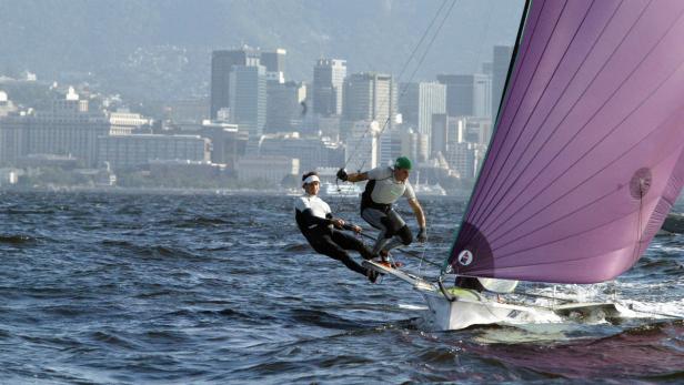 Zwei Segler auf einem Segelboot vor der Skyline einer Stadt.