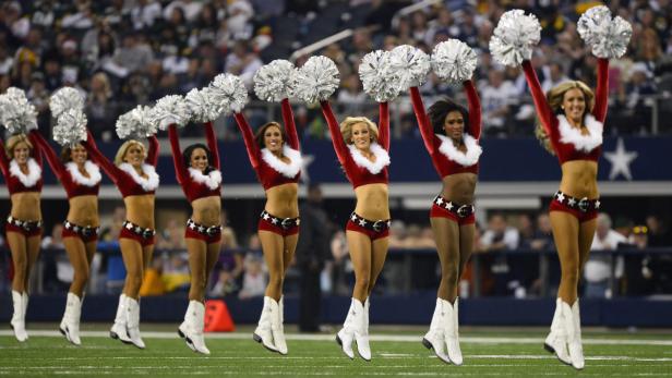 Eine Gruppe Cheerleader in rot-weißen Kostümen springt mit Pompoms auf einem Footballfeld.
