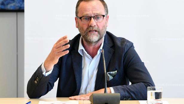 Ein Mann mit Brille gestikuliert an einem Tisch mit Mikrofon und Wasserglas.