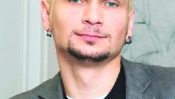 Ein Mann mit blonden Haaren, Sonnenbrille und Ohrringen trägt ein graues Sakko.