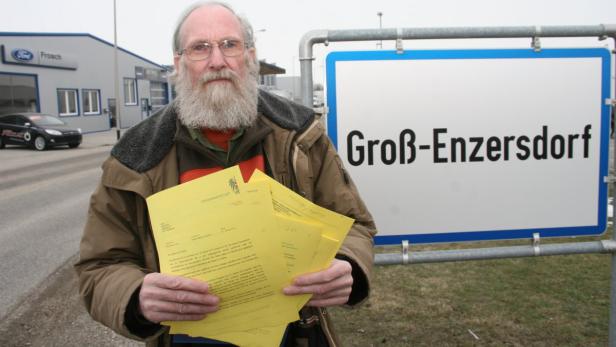 Ein Mann mit Bart steht vor einem Ortsschild von Groß-Enzersdorf und hält gelbe Dokumente in der Hand.
