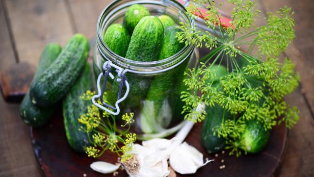 Einmachglas mit Gurken, Dill und Knoblauch auf einem Holzbrett.