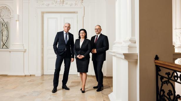 Drei Personen in Anzügen stehen in einem hellen, eleganten Gebäude.