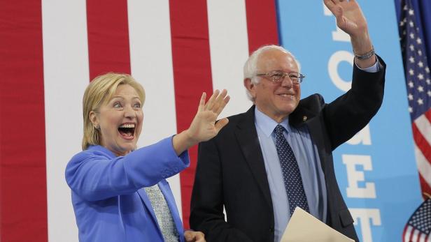 Hillary Clinton und Bernie Sanders winken vor einer amerikanischen Flagge.