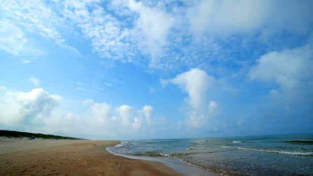 Ein Sandstrand trifft auf das ruhige, blaue Meer unter einem bewölkten Himmel.