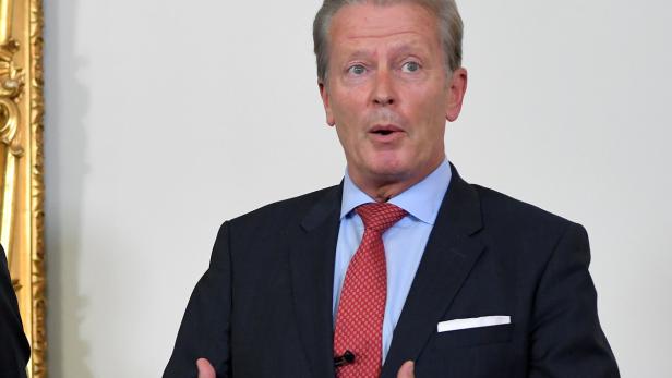 Ein Mann in Anzug und Krawatte gestikuliert vor einer Wand.