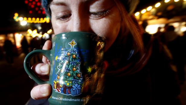 Eine Person trinkt aus einer grünen Tasse mit Weihnachtsbaum-Motiv auf einem festlich beleuchteten Weihnachtsmarkt.