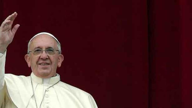 Papst Franziskus winkt vor einem roten Hintergrund.