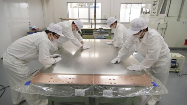 Vier Personen in Reinraumkleidung inspizieren eine große, glänzende Metallplatte.