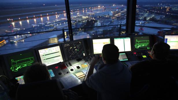 Drei Fluglotsen arbeiten nachts in einem Kontrollturm mit Blick auf einen Flughafen.