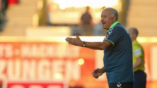 Ein älterer Mann mit Schnurrbart gestikuliert auf einem Sportplatz.