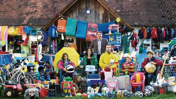 Eine Familie sitzt vor einem Haus, umgeben von zahlreichen Gegenständen für einen Flohmarkt.