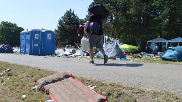 Ein Mann trägt Campingausrüstung auf einem Festivalgelände mit Müll und Zelten.
