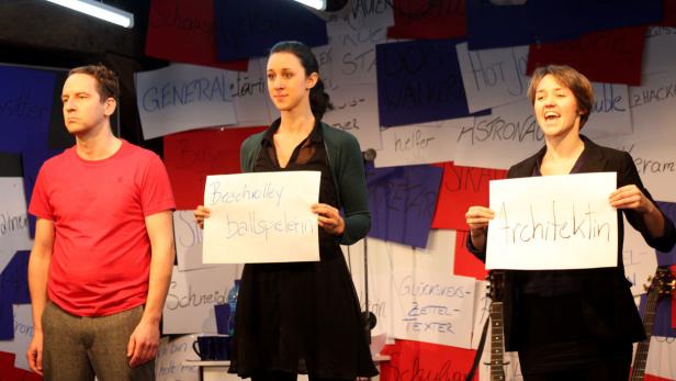 Drei Personen stehen vor einer Wand mit Zetteln und halten Schilder mit Berufsbezeichnungen hoch.