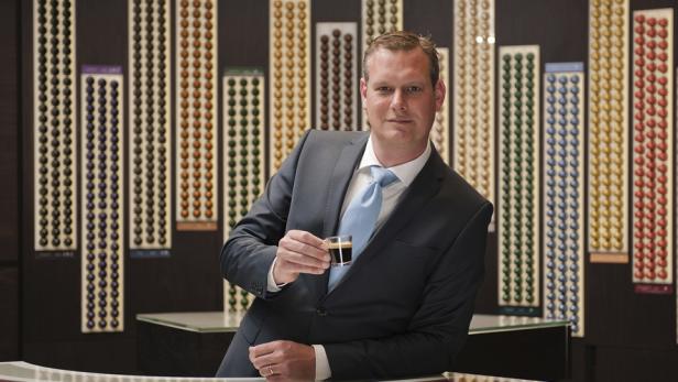 Ein Mann im Anzug hält einen Espresso vor einer Wand mit Kaffeekapseln.