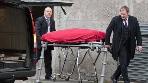 Zwei Männer schieben eine Bahre mit einer roten Decke in einen Leichenwagen.