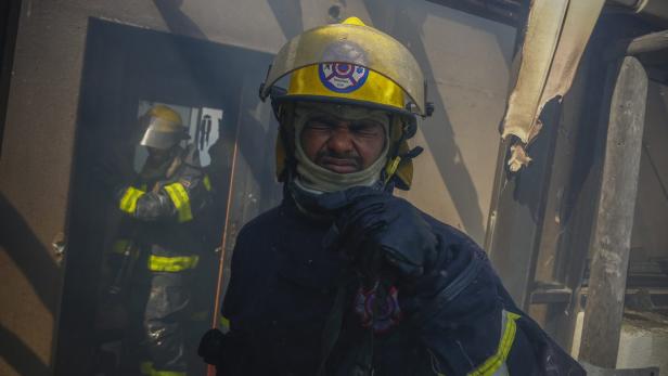 Ein Feuerwehrmann steht inmitten von Rauch und Trümmern, während ein Kollege im Hintergrund zu sehen ist.