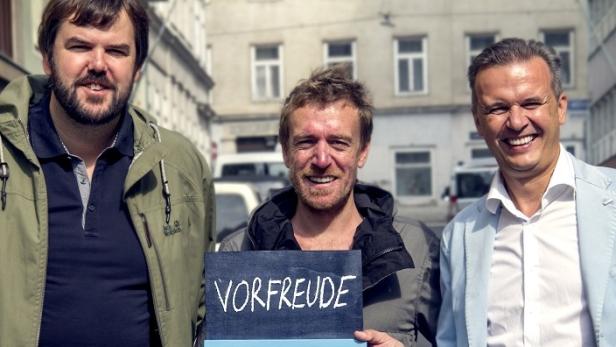 Drei Männer stehen im Freien, einer hält eine Tafel mit der Aufschrift „Vorfreude“.
