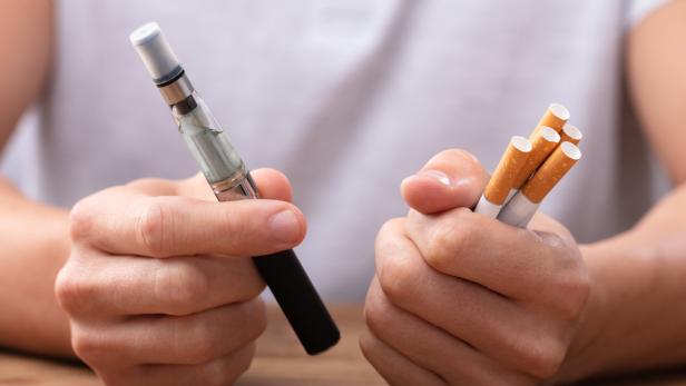 Ein Mann hält eine E-Zigarette in der einen Hand und einige normale Zigaretten in der anderen Hand.