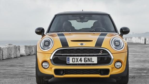 Ein gelber Mini Cooper mit schwarzen Streifen auf der Motorhaube steht auf einer grauen Fläche.
