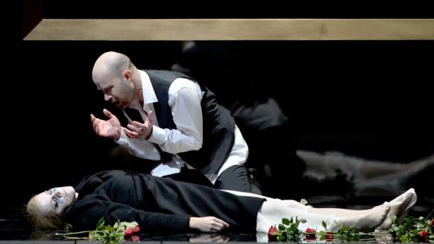 Ein Mann kniet neben einer leblosen Frau, umgeben von roten Rosen.