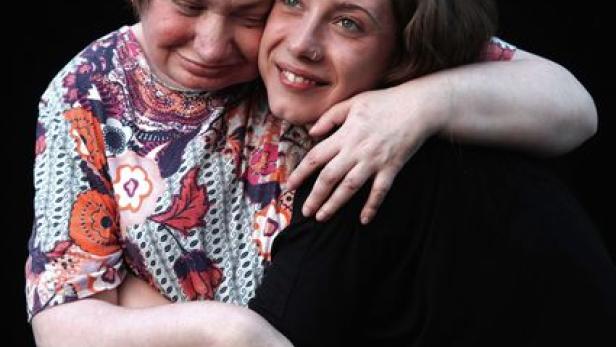 Eine junge Frau umarmt eine ältere Frau vor einem schwarzen Hintergrund.