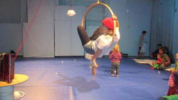 Eine Person mit roter Mütze hängt an einem Trapezring, während Kinder zusehen.