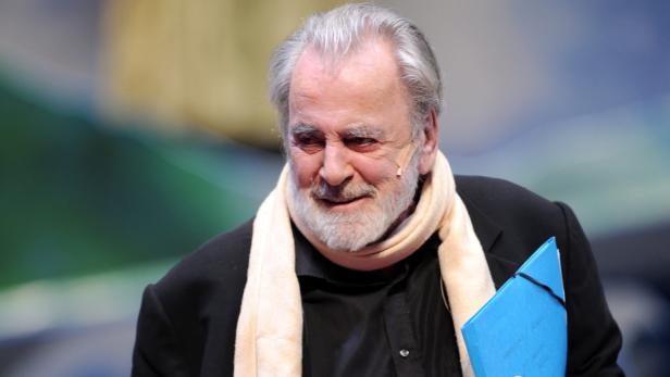 Franco Nero mit Schal und blauem Ordner lächelt.