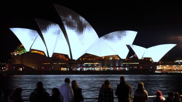 Das Opernhaus von Sydney leuchtet nachts, während Menschen am Wasser stehen.