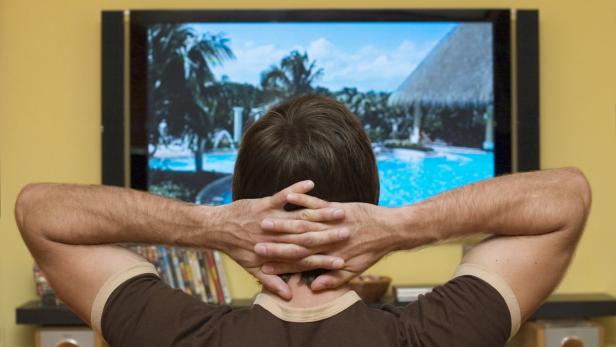 Ein Mann entspannt sich vor dem Fernseher und schaut sich eine Urlaubsszene an.