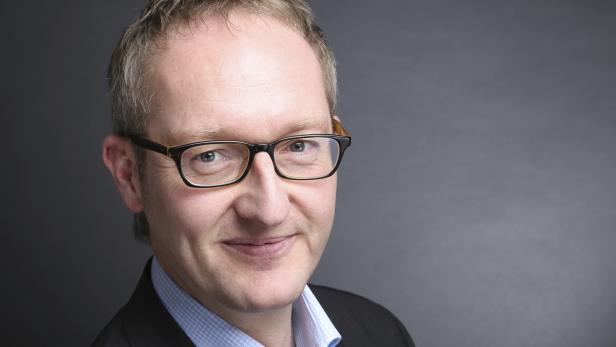 Porträt eines lächelnden Mannes mit Brille vor grauem Hintergrund.
