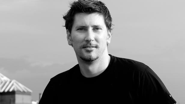 Ein Mann mit Bart und dunklem T-Shirt vor einem grauen Himmel.