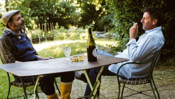 Zwei Männer sitzen an einem Tisch im Garten, einer raucht eine Zigarette.