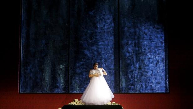 Eine Frau in einem weißen Kleid steht auf einer Bühne vor einem dunklen Hintergrund.