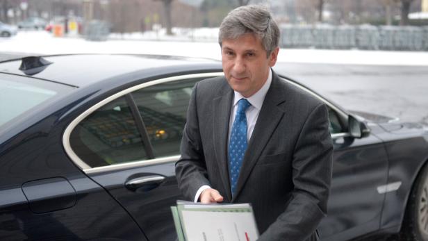 Ein Mann im Anzug steht neben einem dunklen Auto und hält Dokumente in der Hand.