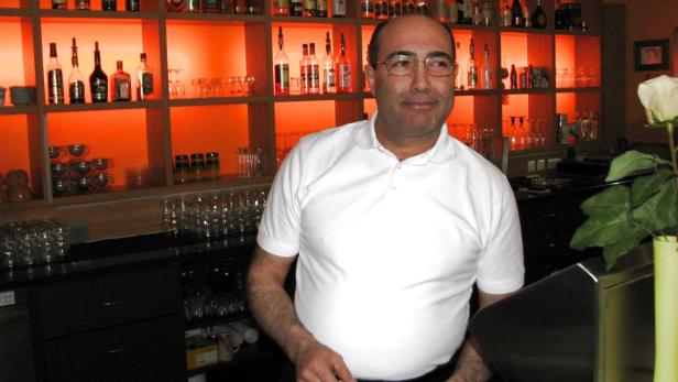 Ein Mann mit Brille steht hinter einer Bar mit Spirituosenregal.