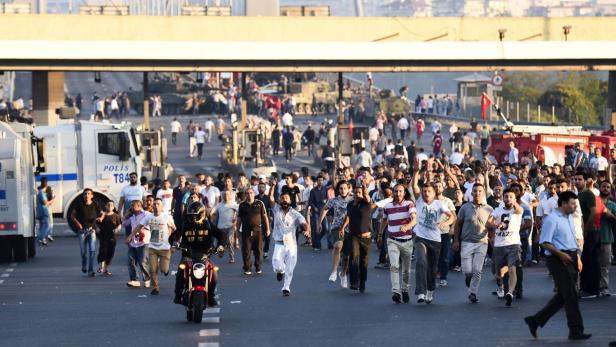 Eine Menschenmenge rennt auf einer Straße an gepanzerten Fahrzeugen und einem Motorradfahrer vorbei.
