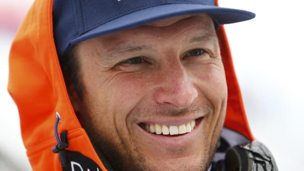 Ein lächelnder Mann mit Bart trägt eine blaue Kappe und eine orangefarbene Jacke mit Kapuze.