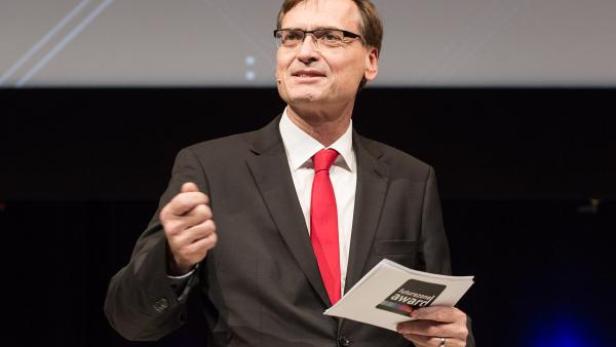 Ein Mann mit Brille und roter Krawatte hält eine Urkunde auf einer Bühne.