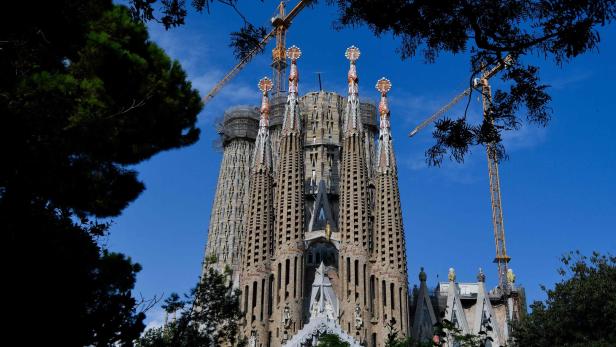 Die Sagrada Familia in Barcelona unter blauem Himmel, teilweise von Bäumen umrahmt.