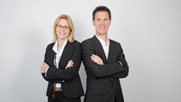 Eine Frau und ein Mann in Anzügen stehen Rücken an Rücken und lächeln.