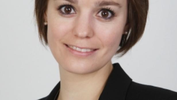 Eine lächelnde Frau mit braunen Haaren und einem schwarzen Blazer vor einem hellen Hintergrund.