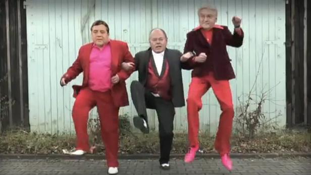 Drei Männer in Anzügen tanzen synchron vor einer grünen Wand.