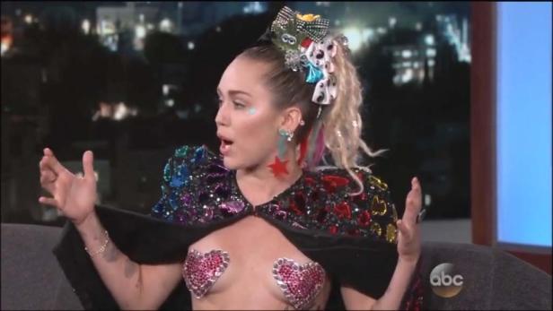 Miley Cyrus gestikuliert während eines Interviews in einem glitzernden Outfit.