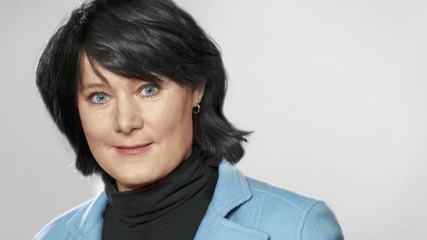 Eine Frau mit blauen Augen und dunklen Haaren trägt ein hellblaues Jackett.