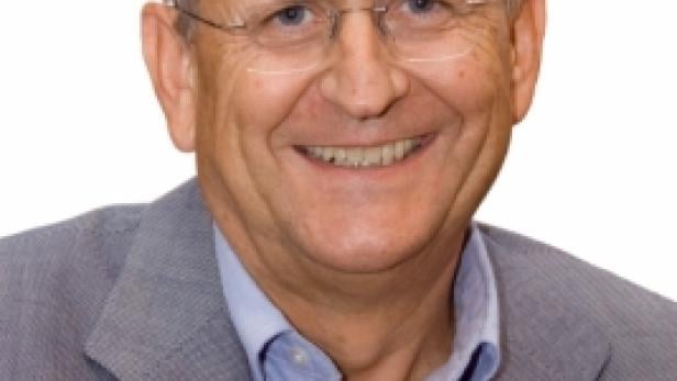 Ein lächelnder älterer Mann mit Brille und grauem Haar trägt ein blaues Hemd und ein graues Jackett.