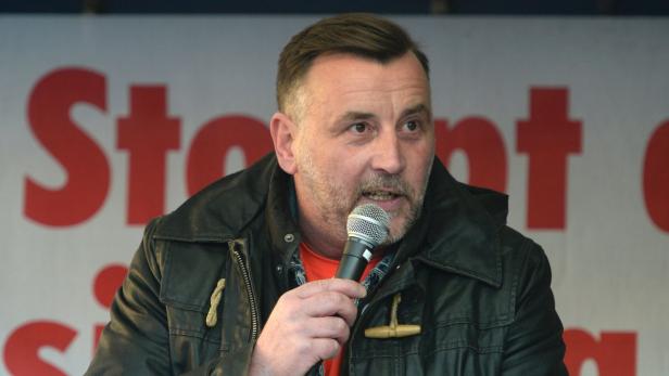 Ein Mann spricht in ein Mikrofon vor einem roten Schriftzug.