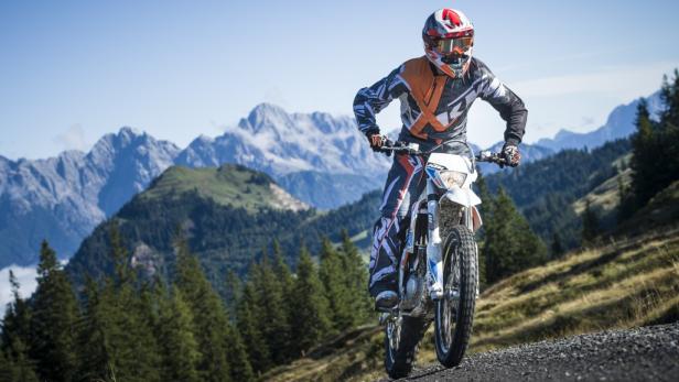 Ein Motocrossfahrer fährt auf einer Schotterpiste vor einer Bergkulisse.