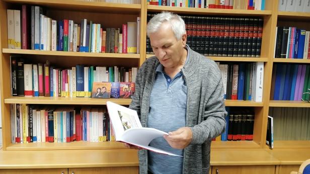 Ein älterer Mann steht vor einem Bücherregal und liest in einem aufgeschlagenen Buch.